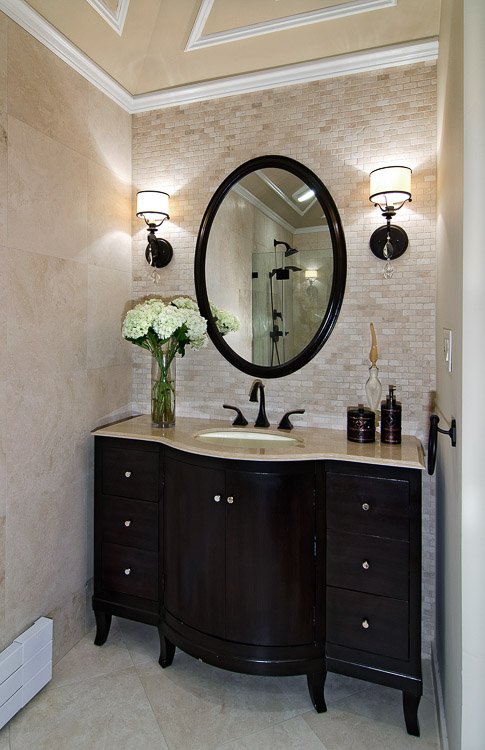 Dark wood bathroom vanity with one sink and a mirror above surrounded with sconces on either side and white flowers on the counter