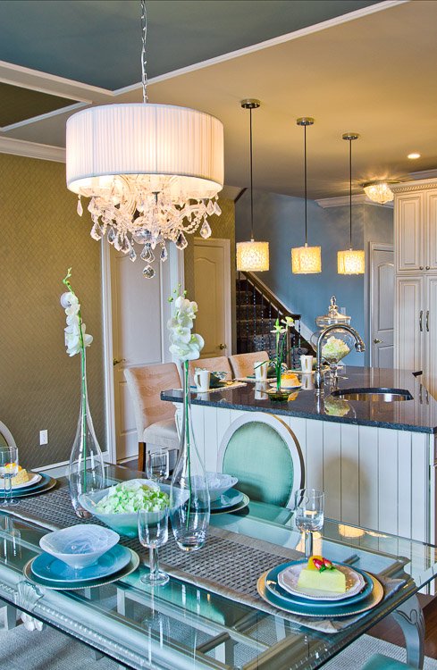 Glass dining table with blue chairs and a chandelier looking into an open concept kitchen