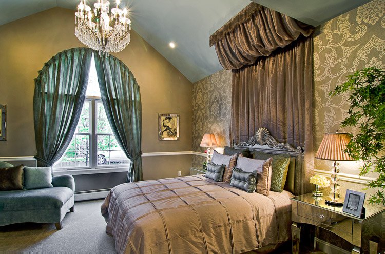 Bedroom with bed with ornate fabric on the wall and ceiling around it, two nighttables with bed side lamps, a large window with curtains, a chaise lounge an a chandelier