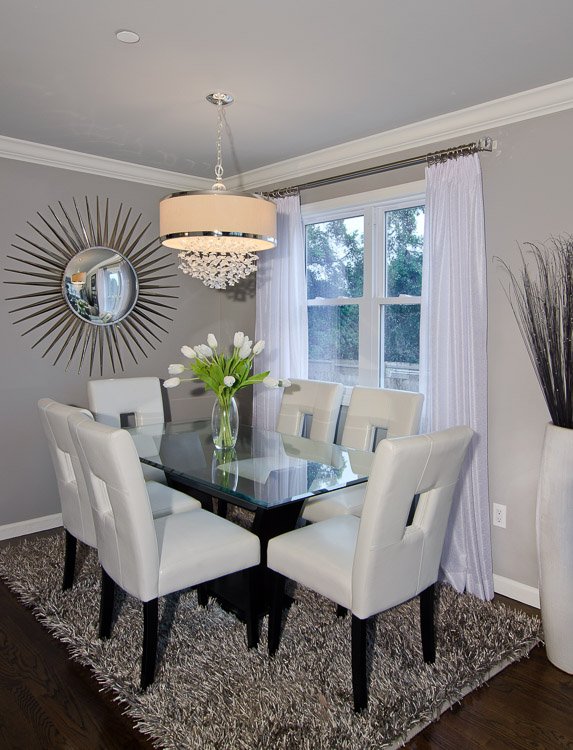 Dining area with a glass table, six white chairs, a chandelier above, a mirror on the wall, a gray shag carpet blow and window with white curtains